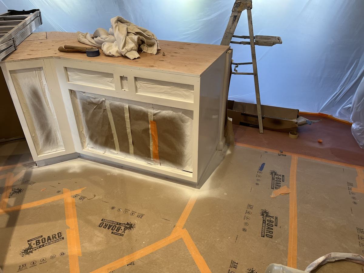 A cabinet carcass standing on a covered floor, primed white, away from the rest of the room.